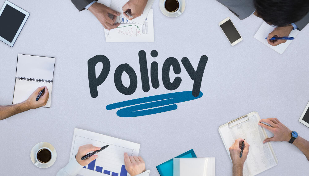 Overhead view of a policy planning meeting: hands write notes and review charts around a table, with 'Policy' and a blue underline in the center.