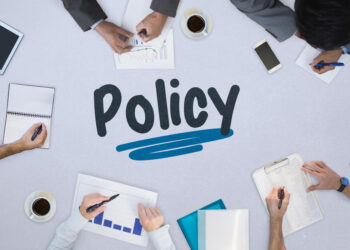 Overhead view of a policy planning meeting: hands write notes and review charts around a table, with 'Policy' and a blue underline in the center.