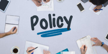 Overhead view of a policy planning meeting: hands write notes and review charts around a table, with 'Policy' and a blue underline in the center.