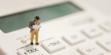 Miniature businessman stands on a calculator key labeled +TAX, looking at papers to symbolize tax calculation.
